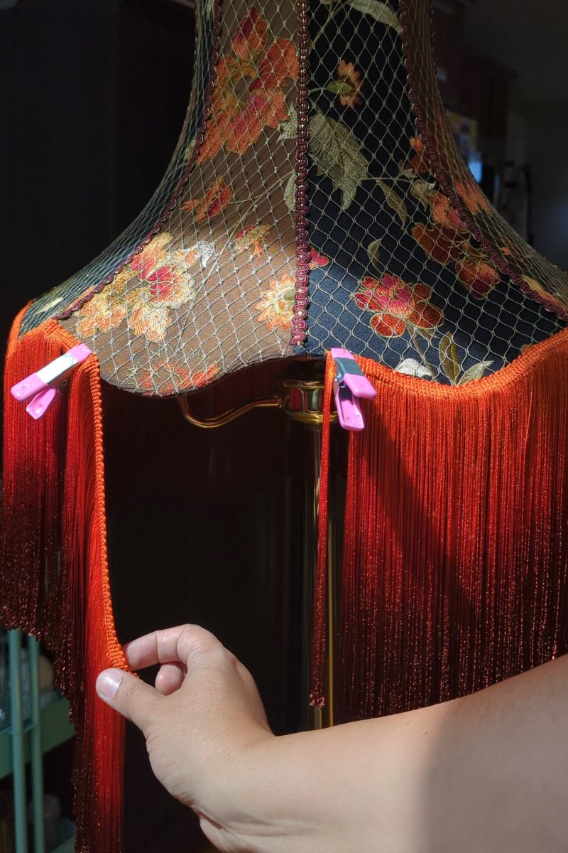 The artist is in the process of applying a fringe to a large lampshade. She is holding the edge of the fringe and pinning it onto the lampshade using clips.