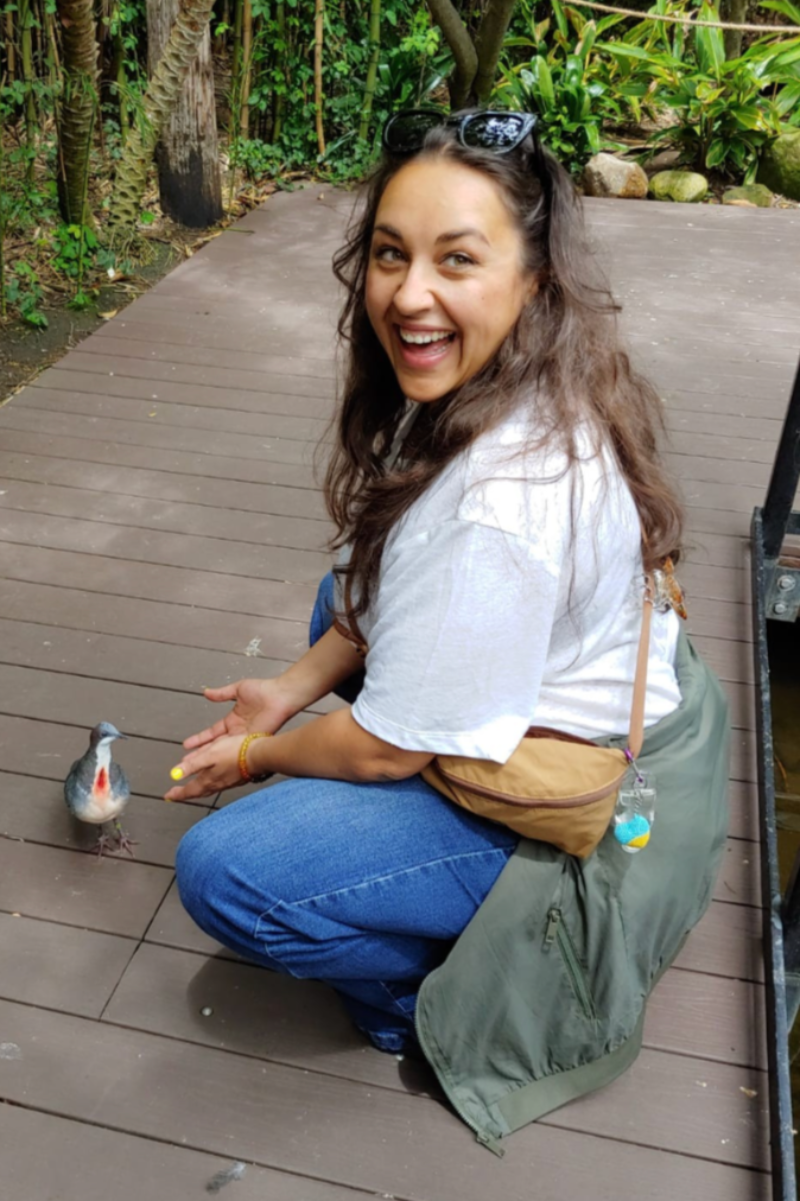 A photo of the artist looking over her shoulder and laughing while squatting next to a small bird with a red breast. 